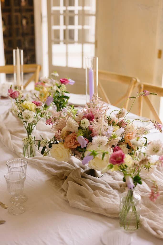 Décoration de table avec un chemin de table rose clair et plusieurs bouquets contenant des fleurs couleur rose et lavande.
