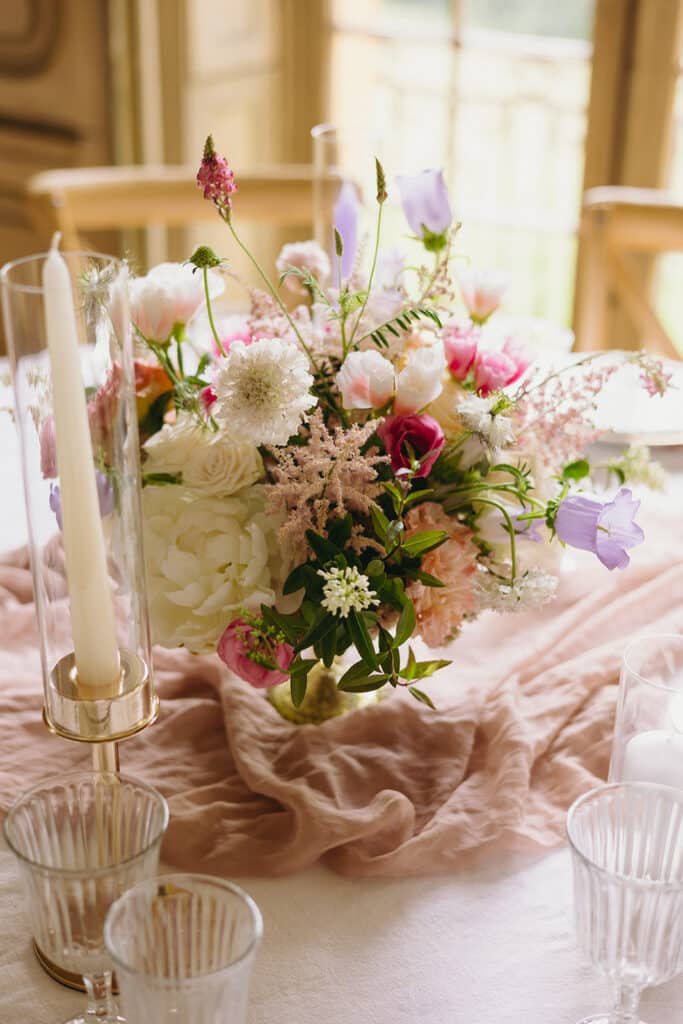 Décoration de table avec un chemin de table rose clair et un bouquet contenant des fleurs couleur rose et lavande dans une coupe sur pied doré