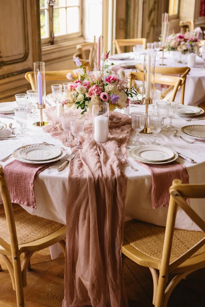 Décoration de table avec un chemin de table rose clair et un bouquet contenant des fleurs couleur rose et lavande dans une coupe sur pied doré